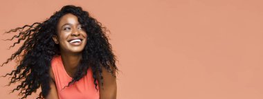 Siyah kıvırcık saçlı, mercan kolsuz gömlek giyen, gülümseyen ve yana bakan siyah bir kadın. Arka plan şeftali renginde. Saçları kabarmış ve doğal bir şekilde hareket ediyor..