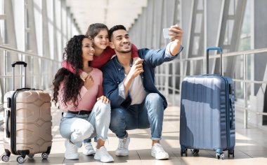 Havalimanı terminalinde Smartphone ile selfie çeken üç kişilik neşeli bir Orta Doğulu ailenin portresi, mutlu Arap ebeveynler ve küçük kızı bavulların yanında otururken kameraya poz veriyorlar.
