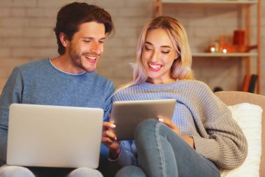 Neşeli eşler dizüstü bilgisayar ve dijital tablet kullanarak evdeki koltukta oturuyorlar. Gadgets Kavramı. Seçici Odaklanma