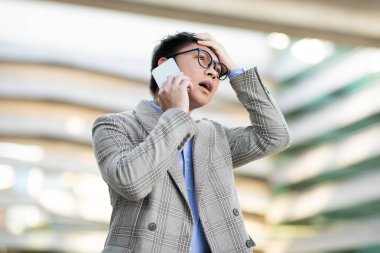 Gözlüklü ve ekose ceketli Asyalı bir adam cep telefonuyla konuşurken kafasını elinde tutuyor. Şaşırmış ve endişeli görünüyor. Dışarıda modern bir binanın önünde duruyor..