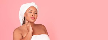 Çekici Afro-Amerikan artı cüsseli kadın yüzüne dokunuyor ve kameraya bakıyor. Besleyici krem kullandıktan sonra ipeksi dokunuşların tadını çıkarıyor. Pembe stüdyo arka planında havluyla poz veriyor.