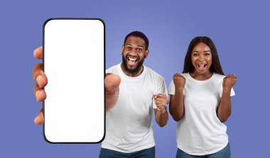 Siyah çift mor bir arka planın önünde duruyor, ellerinde boş ekranlı bir akıllı telefon var. Adam telefonu sol işaret parmağıyla işaret ediyor ve yüzünde kocaman bir gülümseme var..