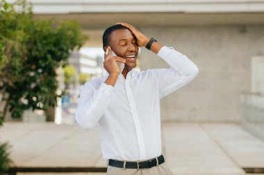 Beyaz gömlek ve pantolon giyen Afro-Amerikalı bir adam binanın dışında duruyor, büyük bir gülümsemeyle telefonuyla konuşuyor. İnançsızlık ya da heyecan içindeymiş gibi elini başının üstüne koydu..