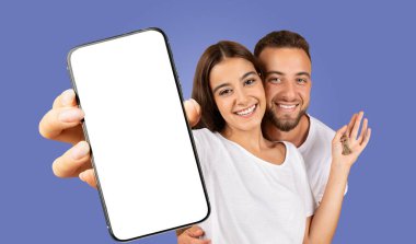 Genç bir çift, saf mavi bir arka planın önünde kameraya parlak bir şekilde gülümsüyor. Kadın elinde boş ekranlı bir akıllı telefon tutuyor.