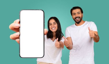 Siyah saçlı, beyaz tişörtlü mutlu bir doğu çifti beyaz ekranlı akıllı telefonu tutarken başparmak kaldırıyor. Çift turkuaz bir arka planın önünde duruyor..
