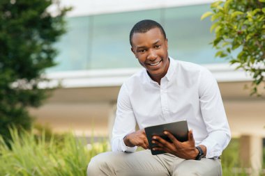 Genç bir Afro-Amerikalı adam dışarıda bir bankta oturuyor, beyaz düğmeli gömlek ve bej pantolon giyiyor ve bir tablet kullanıyor. Gülümsüyor ve doğrudan kameraya bakıyor.