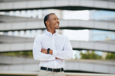 Beyaz gömlek ve haki pantolon giyen Afro-Amerikalı bir adam modern bir binanın önünde kollarını kavuşturmuş duruyor. Yüzünde bir tebessümle yana bakıyor..