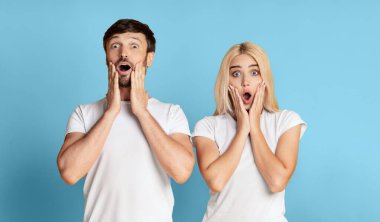 Şaşırmış bir adam ve kadın mavi bir arkaplan önünde duruyor, ikisi de beyaz tişörtler giyiyor ve ağızları açık kameraya bakıyor..