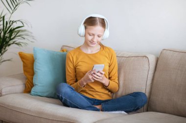 Genç bir kız, sarı gömlek ve kot pantolon giyerek bacak bacak bacak üstüne atmış bir kanepede oturuyor. Kulaklık takıyor ve akıllı telefonunu kullanıyor..