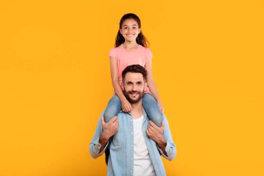 Bir baba ve kızı sarı bir arka planın önünde duruyor. Kız babasının omuzlarında oturuyor, bacakları sarkık. İkisi de gülümsüyor ve kameraya bakıyor..