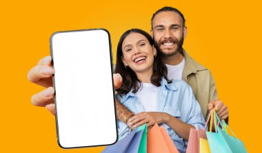 Gülümseyen bir çift canlı sarı bir arka plana karşı duruyor. İkisinde de alışveriş torbası ve boş ekranlı bir akıllı telefon var..