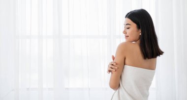 Havlu giymiş neşeli Asyalı kadın pencerenin önünde dikilirken tenine dokunuyor. Beyaz banyo havlusuna sarılı genç bir kadın sabah duşundan sonra yumuşak teninin tadını çıkarıyor.