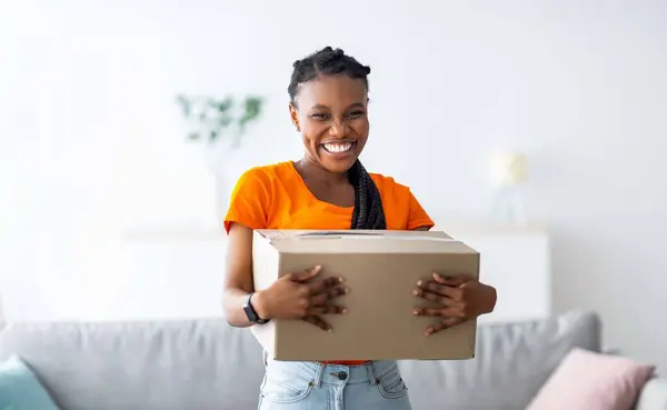 stock image Overjoyed black lady holding cardboard parcel, receiving desired delivery, getting her online order at home. Excited African American woman satisfied with her internet purchase