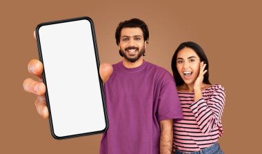 Çok ırklı erkek ve kadın kahverengi bir arka planın önünde duruyor. Adam mor bir tişört giyiyor ve önünde boş bir ekran olan bir akıllı telefon tutuyor..