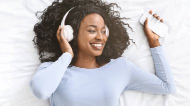 Mobil müzik uygulaması. Mutlu Afrikalı kız yatakta müzik dinliyor, akıllı telefon ve kablosuz kulaklık kullanıyor, fotokopi alanına bakıyor.