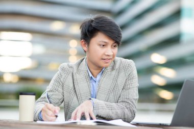 Genç bir Asyalı ofis binasının dışındaki bir masada oturuyor ve dizüstü bilgisayarı üzerinde çalışıyor. O bir kalemle bir deftere yazıyor ve dizüstü bilgisayarı önünde açık..