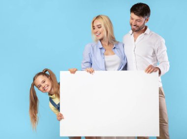Üç kişilik bir aile mavi bir arkaplanın önünde duruyor, ellerinde büyük beyaz bir tabela var. Ebeveynler, bir erkek ve bir kadın, gülümseyin ve tabelaya bakın. Genç kızları yan tarafı gözetliyor.