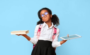 Şirin Afro-Amerikan Küçük Kızı Tablet ve Kitabı Sarı Stüdyo Arkaplanı Üzerine Karşılaştırıyor. Özellikler veya Kitaplar Kavramı.