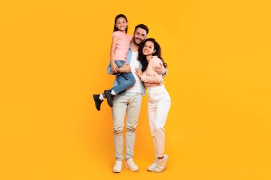 Gülümseyen bir aile, parlak sarı bir arka plana karşı birlikte durur. Baba kızını kollarında tutarken, anne onun yanında, kolunu beline dolamış.