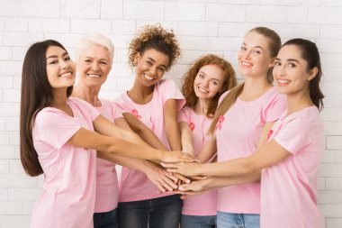 Kanser ve Destek. Gülümseyen Karışık Kadın Gönüllüler Farkındalık Tişörtleri Pembe Kurdeleli El ele tutuşan Kapalı Beyaz Duvarın yanında