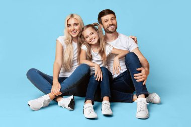 Genç bir aile, bir anne, bir baba ve bir kız, açık mavi bir arka planda otururken gülümsüyor ve kameraya bakıyor. Beyaz tişört ve kot pantolon giyiyorlar.