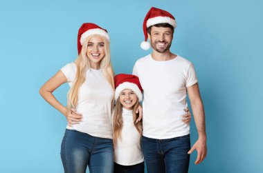 Kırmızı Noel Baba şapkası takan üç kişilik gülümseyen bir aile mavi arka planda duruyor. Ebeveynler çocuklarının her iki yanında, elleri birbirlerinin belinde dururlar..