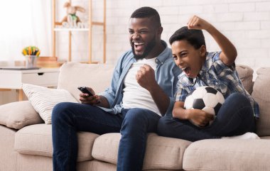 Afro baba ve oğul futbol fanatikleri evde futbol izleyip duygusal tezahüratlar yapıyor, futbol topuyla oturma odasında kanepede oturuyor.