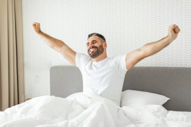 Beyaz tişörtlü bir adam yatakta oturuyor, kollarını yana doğru açıyor ve gülümsüyor. O, gerilirken tavana doğru bakıyor. Yatağın beyaz çarşafları ve gri bir yatak başlığı var.