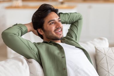 Sakallı bir Arap beyaz bir kanepede oturuyor, kolları başının arkasında, rahat ve hoşnut görünüyor. Arka plan oturma odası gibi görünüyor..