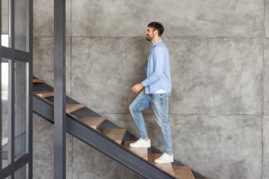 Mavi gömlekli ve kot pantolonlu bir adam modern bir iç merdivende tahta merdivenler ve siyah metal parmaklıklarla yürüyor. Beton bir duvara dayanan merdivenleri çıkarken gülümsüyor..