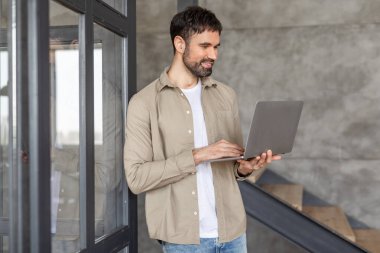 Sakallı bir adam modern bir ofiste cam bir kapının yanında duruyor ve bir dizüstü bilgisayar üzerinde çalışıyor. Beyaz bir tişört ve kot pantolonun üzerine açık kahverengi bir gömlek giyiyor. Gülümsüyor ve odaklanmış görünüyor.