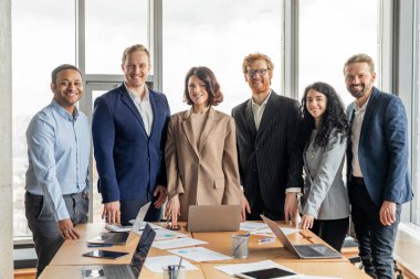 Bir grup farklı iş adamları modern bir ofis alanında durup fotoğraf için gülümsüyorlar. Takım hem erkeklerden hem de kadınlardan oluşur. İş kıyafeti giymişler.