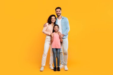 Üç kişilik mutlu bir aile, parlak sarı bir arka plana karşı poz veriyor. Baba, anne ve kız birbirlerine yakın dururlar, ebeveynler genç kıza sarılırlar..