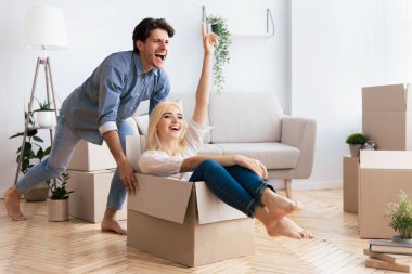 Bir kadın karton bir kutuda otururken, bir adam onu yeni bir eve itiyor, ikisi de gülüyor..