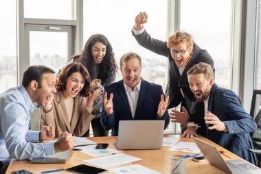 Aralarında kadın ve erkeklerin de yer aldığı çeşitli iş arkadaşları, modern bir ofiste dizüstü bilgisayarın etrafında toplanıyorlar. Hepsi heyecanla gülümsüyor ve gülüyor, birkaç kolu havada..