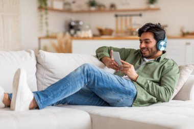 Arap adam beyaz bir kanepede yatıyor, kot pantolon ve yeşil gömlek giyiyor. Kulaklığı var ve gülümseyerek akıllı telefonuna bakıyor. Rahatlıyor ve eğleniyor gibi görünüyor..