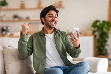 Arap adam evdeki bir kanepede oturmuş, akıllı telefonuna heyecanla bakıyor. Gülümsüyor ve yumruğunu havaya kaldırıyor. Başarılı bir sonucu kutluyor.