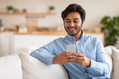 Arap adam beyaz bir kanepede oturur ve yüzünde bir gülümsemeyle telefonuna bakar. Adam mavi düğmeli bir gömlek giyiyor. Arka plan bulanık ve bir mutfak alanı gösteriyor..