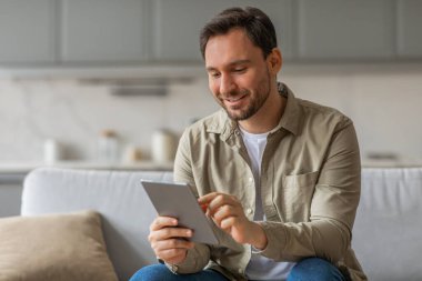 Oturma odasındaki koltukta oturan bir adam tablet bilgisayar kullanırken gülümsüyor. Bej düğmeli gömlek ve kot pantolon giyiyor..