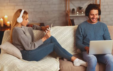 Evde Kanepede Rahatlatıcı Kulaklık Giyen Laptop ve Tablet Kullanan Pozitif Eşler. Modern Yaşam Tarzı Kavramı.