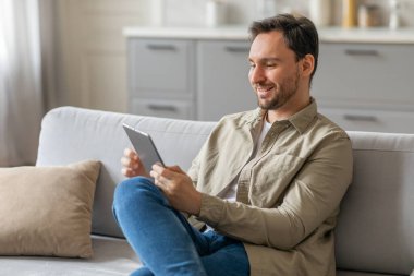 Bronz gömlekli ve mavi kotlu bir adam açık gri bir koltukta oturuyor, iki elinde de tablet var. O, bir gülümsemeyle tablete bakar. Kanepe modern bir oturma odasında, beyaz, modern bir mutfağı var.