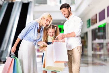 Genç bir kız anne ve babasının arasında duruyor, üçü de alışveriş poşetlerine bakıyor. Kız gülümsüyor ve ailesi çantaların içindekilere bakarken gülümsüyor..