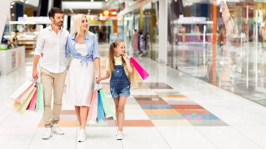 Üç kişilik mutlu bir aile, alışveriş merkezlerinde ellerinde alışveriş torbalarıyla dolaşıyorlar. Kızları mavi kot pantolon ve pembe gömlek giyiyor..