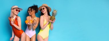 Mayo giymiş üç kadın ellerinde meyvelerle parlak mavi bir arka planda duruyorlar. Bir kadın bir ananas tutuyor, diğeri iki portakal tutuyor, ve üçüncü kadın da bir muz tutuyor, kopyalama alanı.