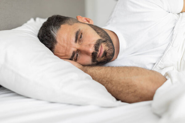 A man is lying in bed with his arm under his head. He is resting his head on a white pillow, and he has a small beard. The man appears to be sleepless, suffering from depression