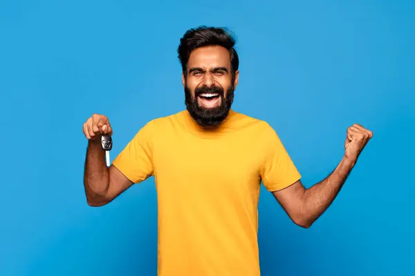 stock image Hindu man with a beard is smiling and holding up a car key in his right hand while his left fist is clenched in the air. He is wearing a yellow t-shirt and is standing in front of a blue background.