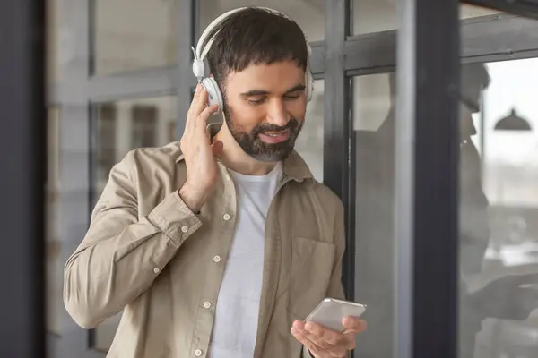 stock image A man with a beard and short hair, wearing a beige button-down shirt and white undershirt, adjusts his headphones while holding a smartphone. He is standing in a modern office building