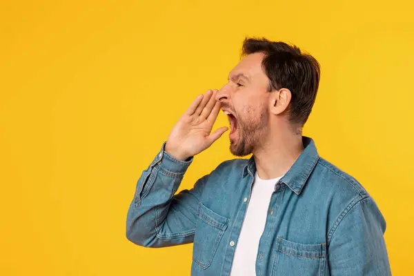 stock image A man with short, dark hair, wearing a light blue button-up shirt, is yelling with his mouth open and a hand cupped near his mouth. He is standing against a plain, bright yellow background.