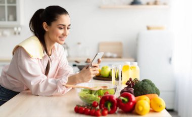Mutfakta yemek pişirirken cep telefonu kullanan gülümseyen genç bir kadın masaya yaslanan neşeli bir kadın cep telefonundan mesajlar okuyan ve gülümseyen, bedava fotokopi çeken bir kadın.