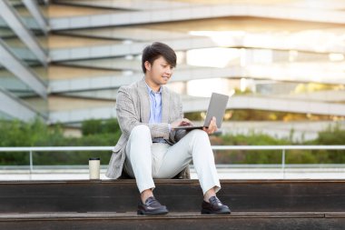 Spor ceket ve beyaz pantolon giymiş genç bir Asyalı modern bir binanın önünde dizüstü bilgisayarıyla çalışırken beton bir basamakta oturuyor..
